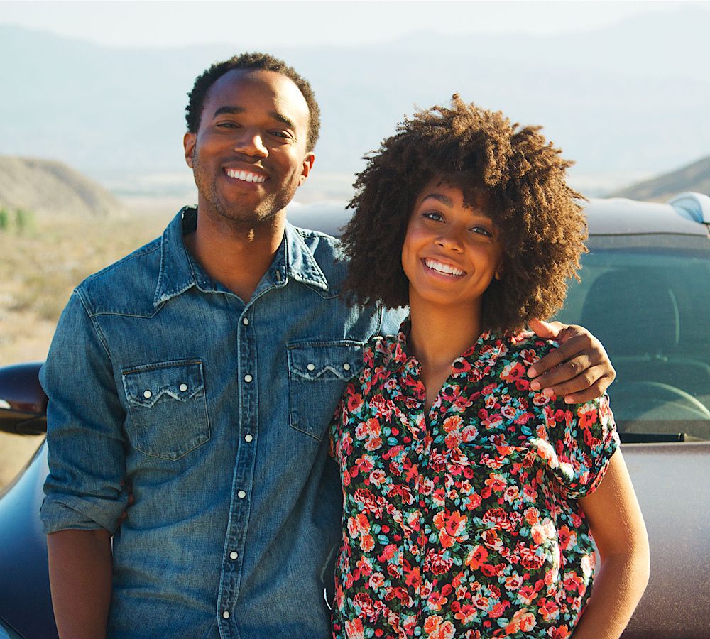Portrait Of Smiling Young Couple On Road Trip Standing By Rental Car On Country Road by monkeybusiness